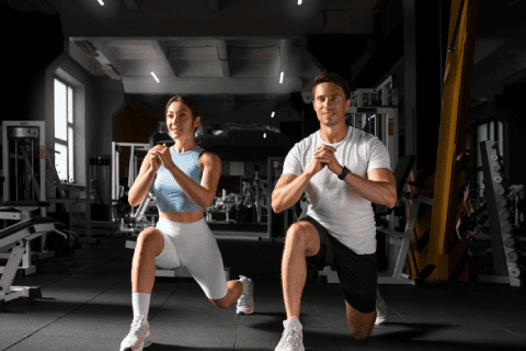 Fitness Center Two people are exercising in a gym, performing lunges side by side. The woman on the left wears a light blue tank top and white shorts, while the man on the right wears a white t-shirt and black shorts. Both are focused on their workout. Equipment is visible in the background.