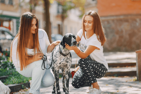 pet friendly community Two young women are outdoors with a black and white dog. One woman, wearing white pants and a white top, crouches while the other, wearing a white top and patterned leggings, pets the dog. They are on a paved path with greenery and buildings in the background.
